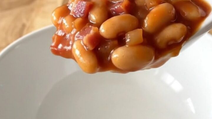 A white bowl filled with slow-cooked baked beans, featuring visible pieces of bacon, onion, and a thick, glossy tomato sauce.
