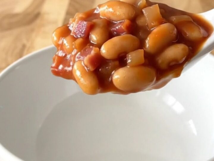 A white bowl filled with slow-cooked baked beans, featuring visible pieces of bacon, onion, and a thick, glossy tomato sauce.