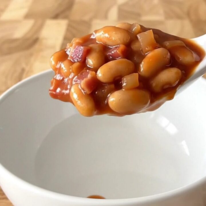 A white bowl filled with slow-cooked baked beans, featuring visible pieces of bacon, onion, and a thick, glossy tomato sauce.