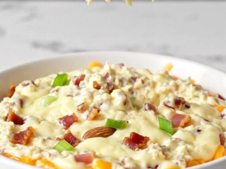 A close-up of a creamy million dollar dip in a white bowl, topped with bacon crumbles, almonds, and green onions.