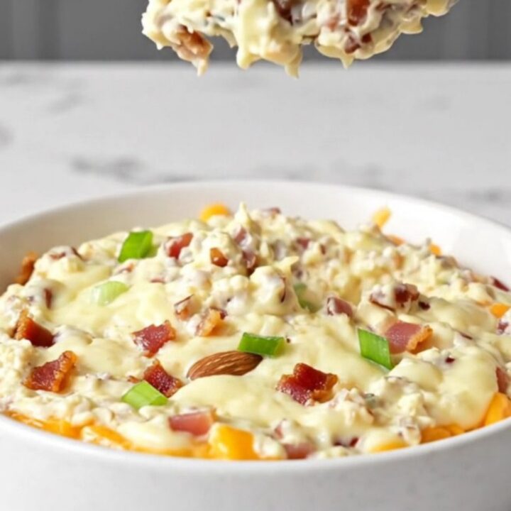 A close-up of a creamy million dollar dip in a white bowl, topped with bacon crumbles, almonds, and green onions.