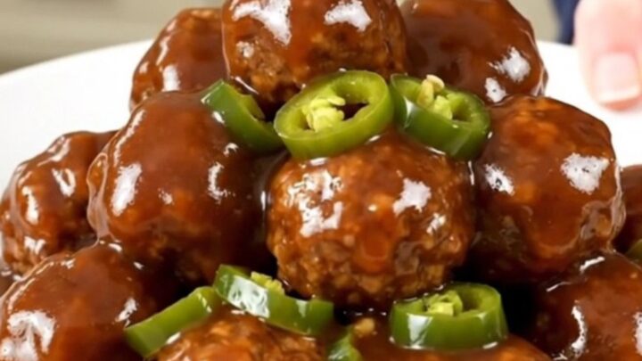 A close-up of slow cooker meatballs coated in a glossy, dark barbecue sauce with visible candied jalapeno slices.