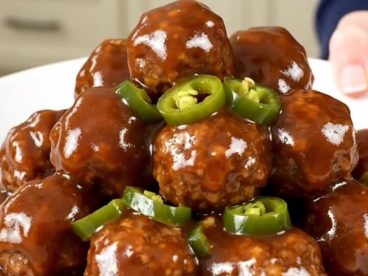 A close-up of slow cooker meatballs coated in a glossy, dark barbecue sauce with visible candied jalapeno slices.