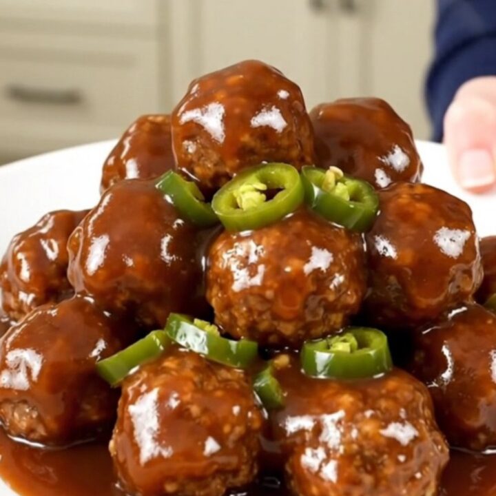 A close-up of slow cooker meatballs coated in a glossy, dark barbecue sauce with visible candied jalapeno slices.