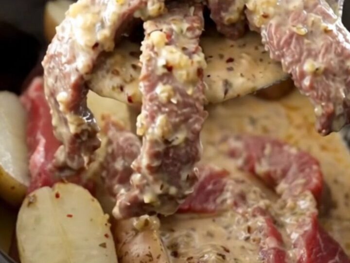 A close-up of tender round steak and baby red potatoes in a creamy garlic Parmesan sauce inside a slow cooker.