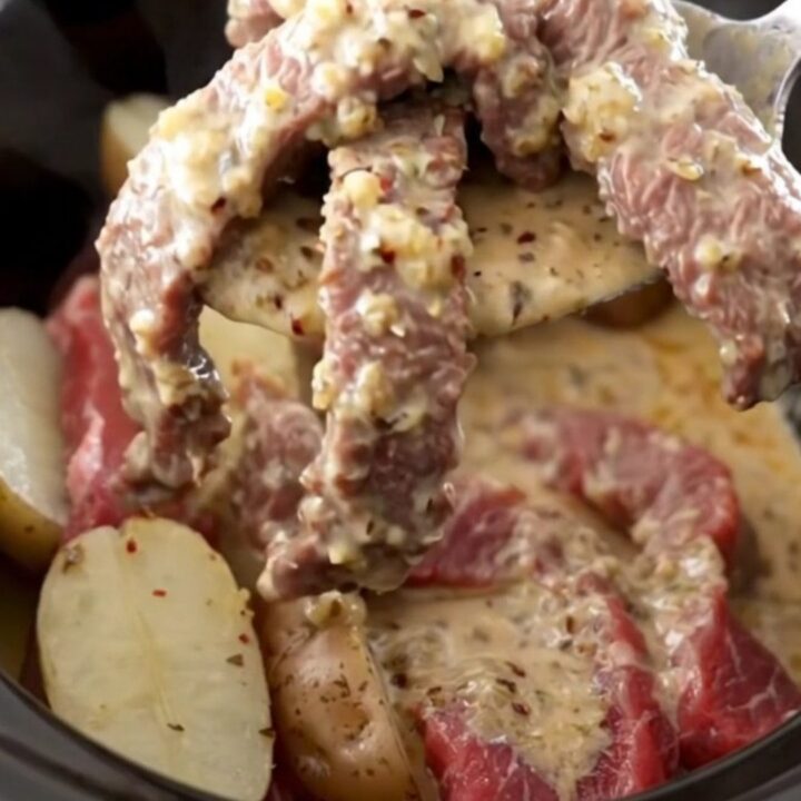 A close-up of tender round steak and baby red potatoes in a creamy garlic Parmesan sauce inside a slow cooker.
