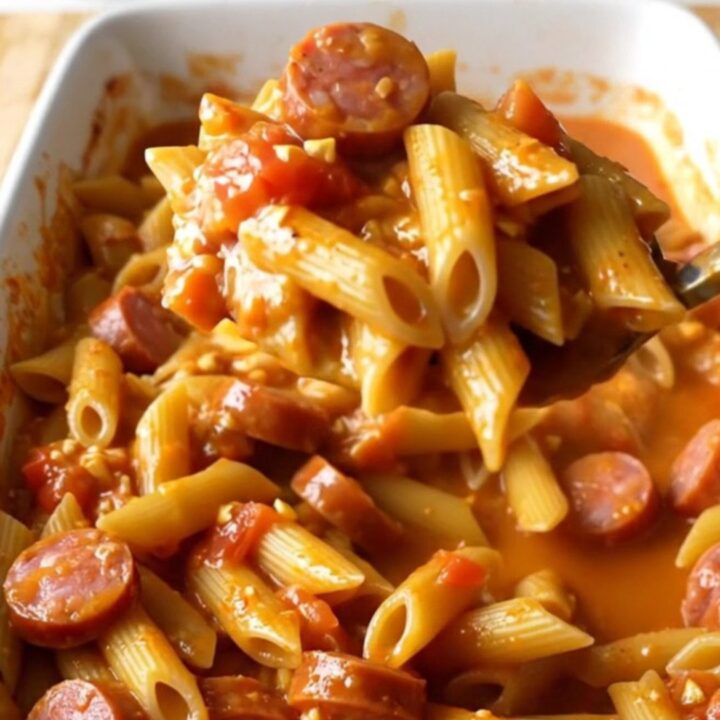A top-down view of a rectangular white baking dish filled with penne pasta, sliced smoked sausage, diced tomatoes