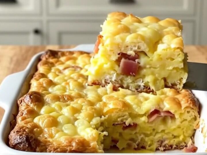 A golden-brown eggs Benedict breakfast casserole in a white ceramic dish on a light wooden counter.