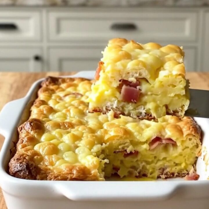 A golden-brown eggs Benedict breakfast casserole in a white ceramic dish on a light wooden counter.