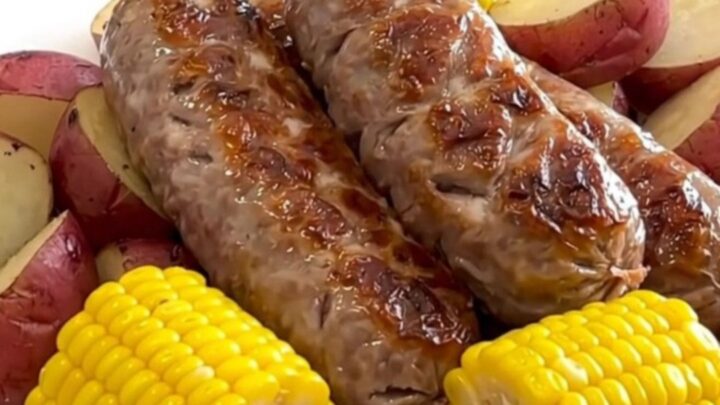 A close-up of a white plate holding a juicy bratwurst link, a piece of corn on the cob, and halved red potatoes.