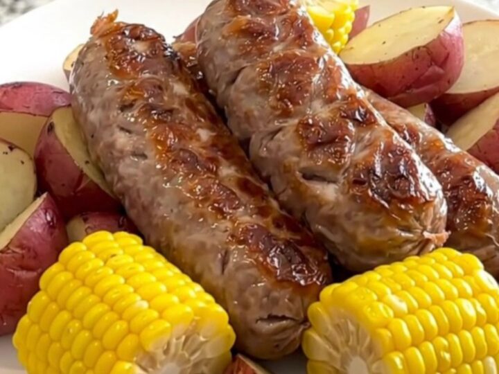 A close-up of a white plate holding a juicy bratwurst link, a piece of corn on the cob, and halved red potatoes.