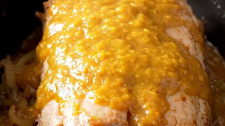A whole, tender slow-cooked pork loin glazed in a golden apricot Dijon sauce with onions in a black slow cooker.