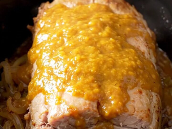 A whole, tender slow-cooked pork loin glazed in a golden apricot Dijon sauce with onions in a black slow cooker.