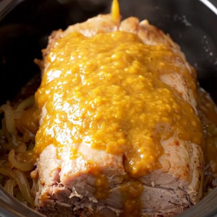 A whole, tender slow-cooked pork loin glazed in a golden apricot Dijon sauce with onions in a black slow cooker.