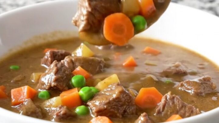 A close-up of a white bowl filled with creamy beef pot pie stew featuring tender beef cubes, potatoes, peas, and carrots.