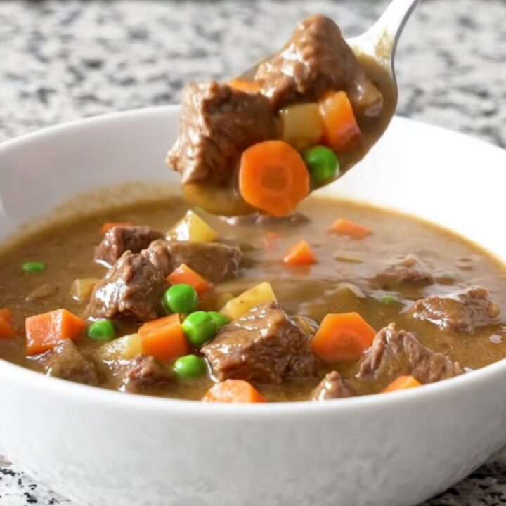 A close-up of a white bowl filled with creamy beef pot pie stew featuring tender beef cubes, potatoes, peas, and carrots.