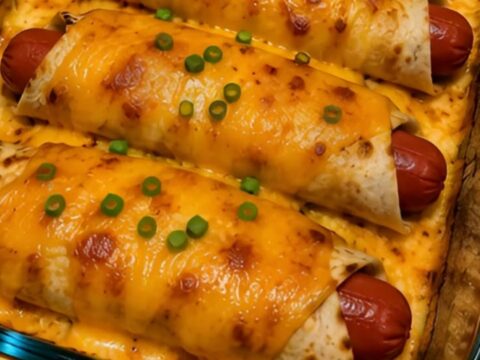 Four hot dog cheddar wraps in a glass baking dish, golden-brown, bubbly, and garnished with chopped green onions.