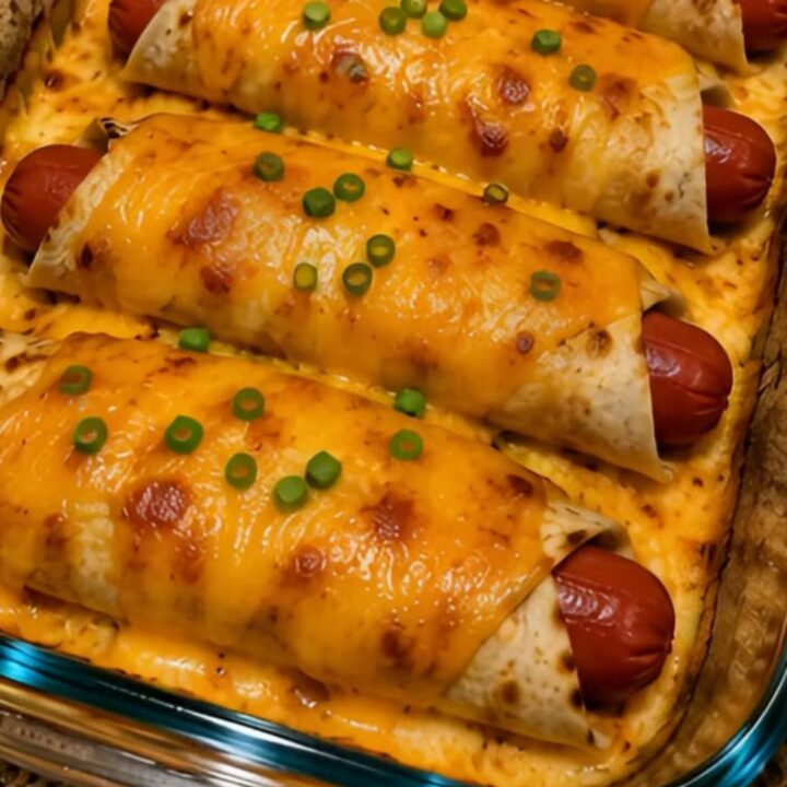 Four hot dog cheddar wraps in a glass baking dish, golden-brown, bubbly, and garnished with chopped green onions.