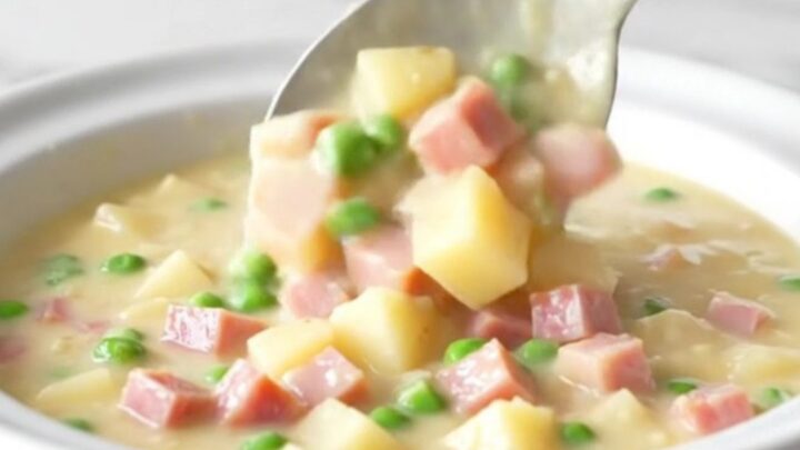A bowl of creamy ham and potato chowder with visible cubes of ham, tender gold potatoes, and bright green peas.