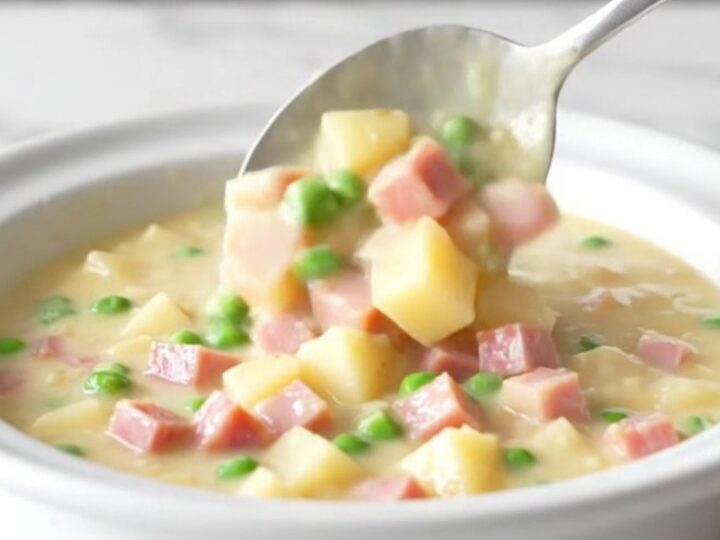 A bowl of creamy ham and potato chowder with visible cubes of ham, tender gold potatoes, and bright green peas.