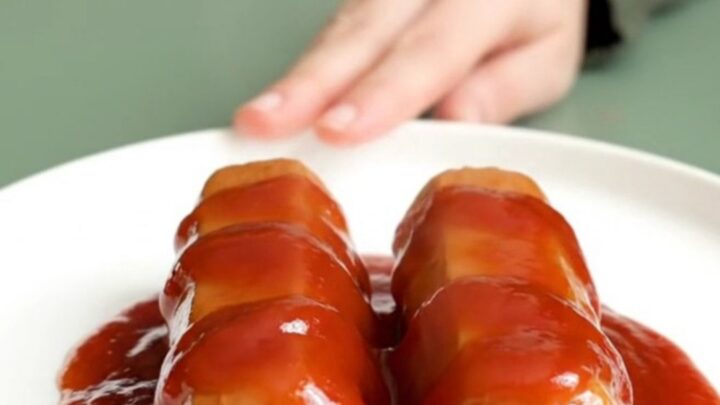 A close-up view of bite-sized hot dog pieces coated in a glossy, thick, red bourbon glaze, served on a white plate.