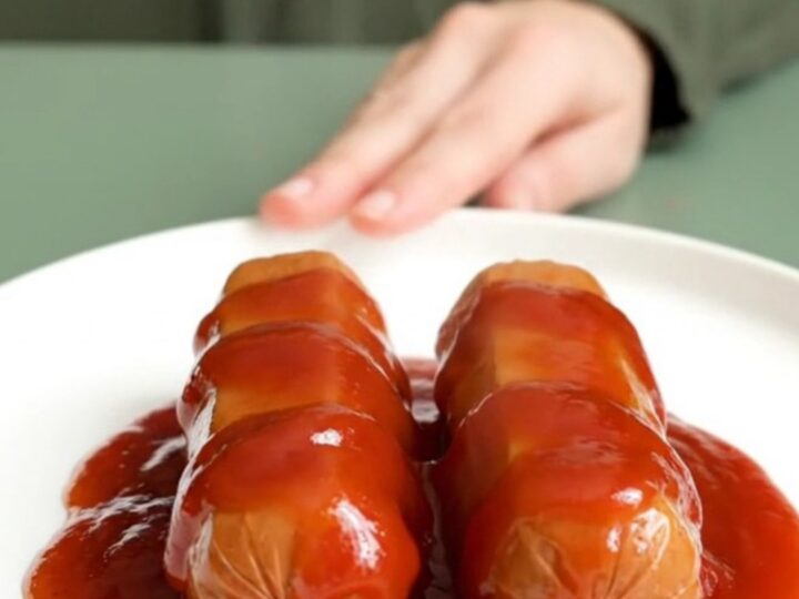 A close-up view of bite-sized hot dog pieces coated in a glossy, thick, red bourbon glaze, served on a white plate.