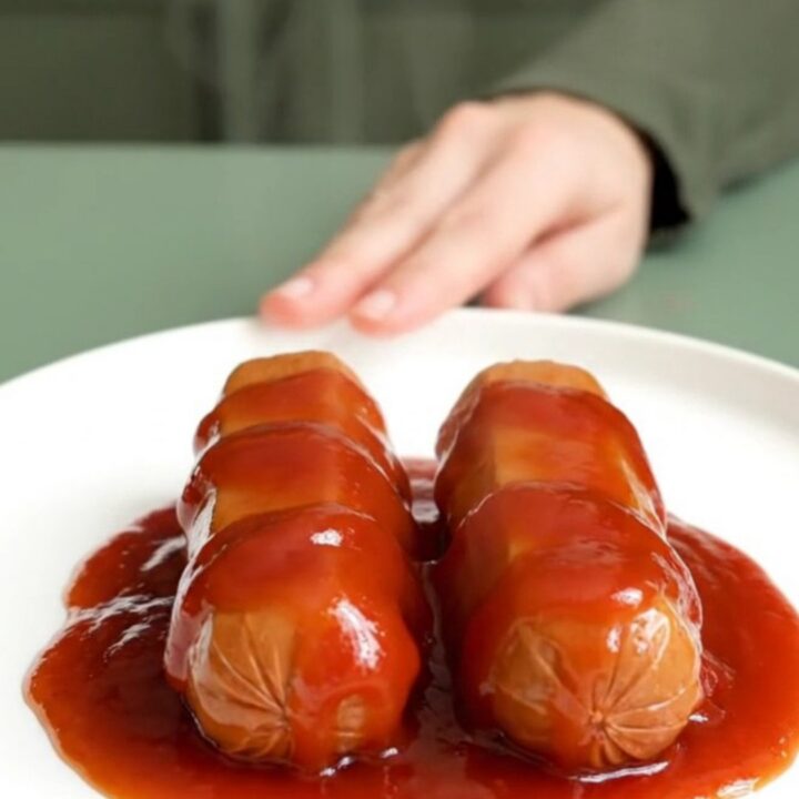 A close-up view of bite-sized hot dog pieces coated in a glossy, thick, red bourbon glaze, served on a white plate.