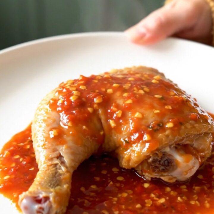A close-up of tender, slow-cooked chicken leg quarters coated in a thick, glossy chili garlic sauce on a white plate.