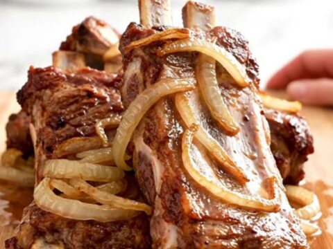 A close-up of tender, slow-cooked spareribs covered in a tangy vinegar sauce with soft onions on a wooden board.