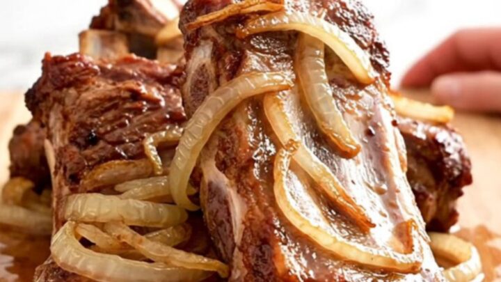 A close-up of tender, slow-cooked spareribs covered in a tangy vinegar sauce with soft onions on a wooden board.
