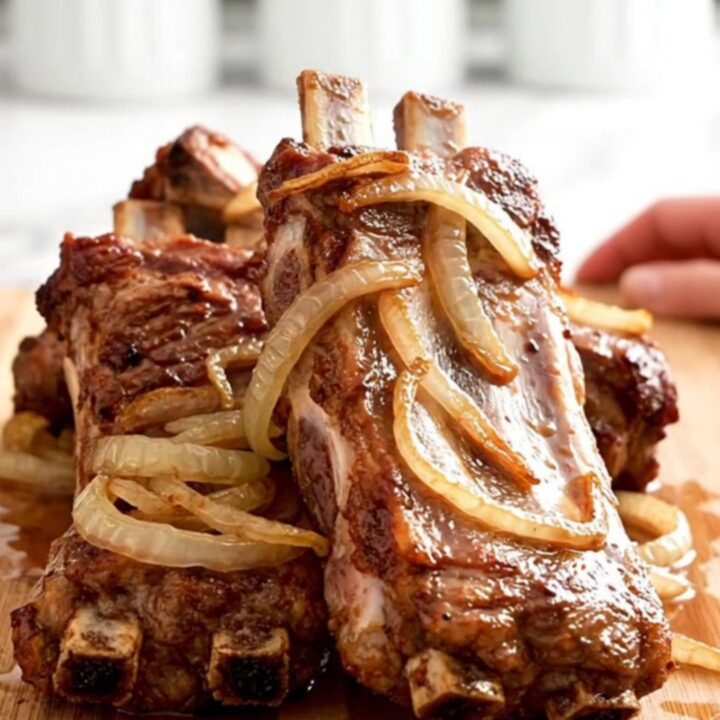 A close-up of tender, slow-cooked spareribs covered in a tangy vinegar sauce with soft onions on a wooden board.