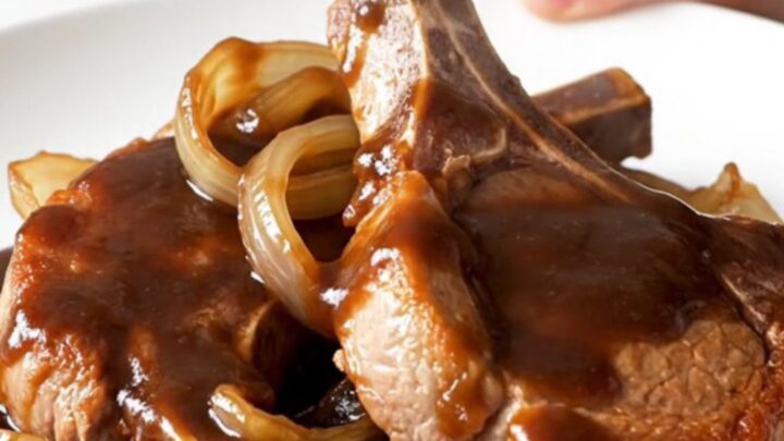 A close-up of tender, bone-in pork chops glazed in a dark, glossy root beer barbecue sauce with soft onions.