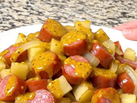 A close-up of slow-cooked sausage, gold potato cubes, and onions coated in a yellow mustard barbecue sauce.
