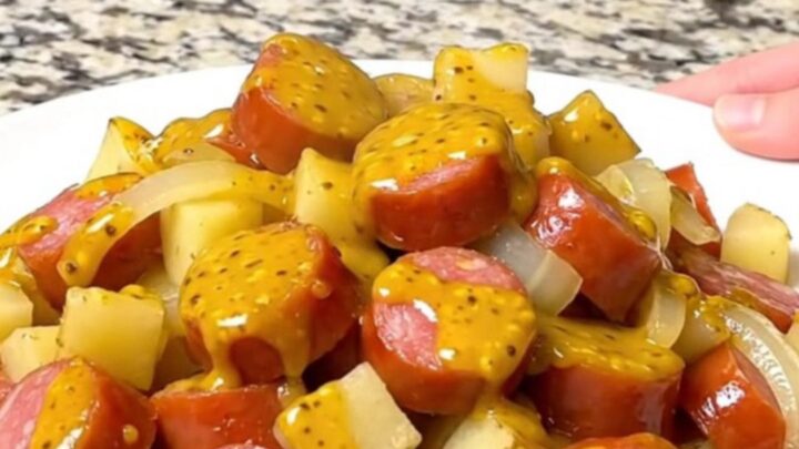 A close-up of slow-cooked sausage, gold potato cubes, and onions coated in a yellow mustard barbecue sauce.