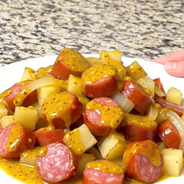 A close-up of slow-cooked sausage, gold potato cubes, and onions coated in a yellow mustard barbecue sauce.