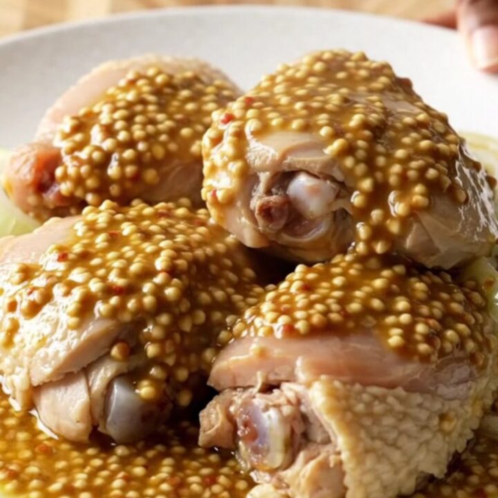 A close-up of tender, braised chicken thighs topped with soft, caramelized onion slices in a rich mustard sauce.