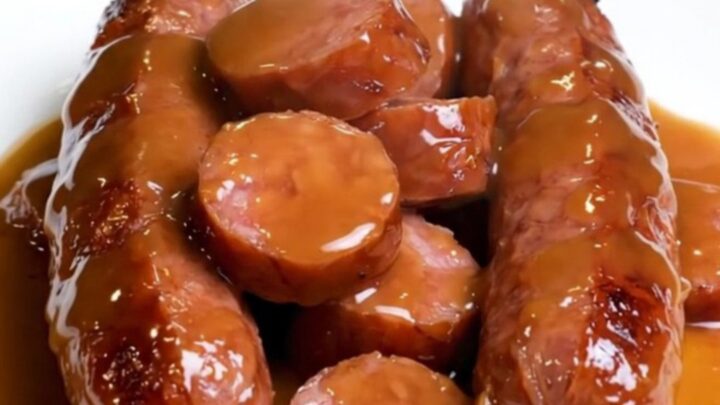 A close-up of slow-cooked kielbasa coins and baby carrots glazed in a glossy brown sugar sauce.
