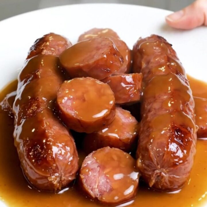 A close-up of slow-cooked kielbasa coins and baby carrots glazed in a glossy brown sugar sauce.