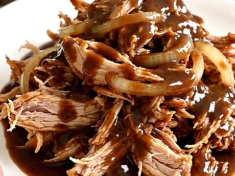 A slow cooker filled with tender shredded chicken breasts coated in a rich, dark root beer sauce and onion slices.