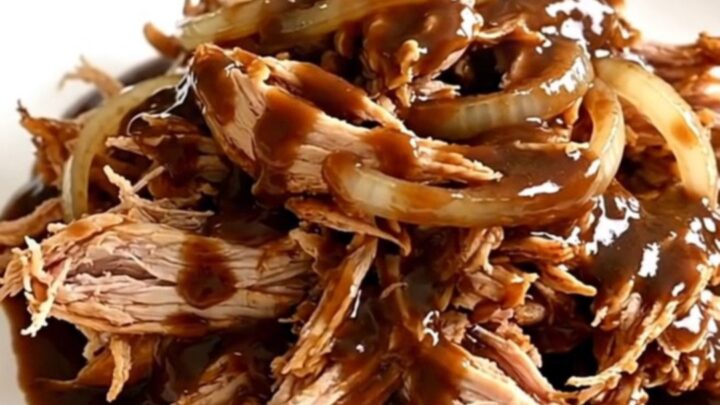 A slow cooker filled with tender shredded chicken breasts coated in a rich, dark root beer sauce and onion slices.