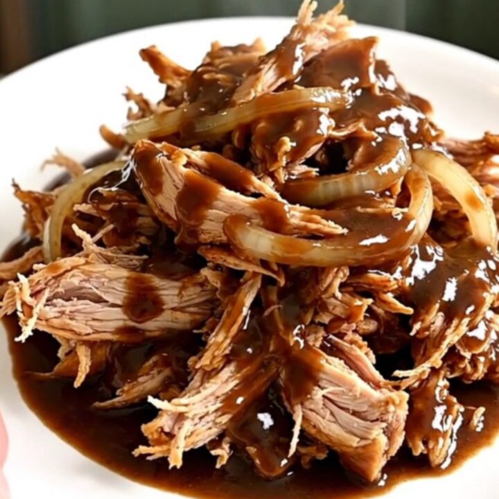 A slow cooker filled with tender shredded chicken breasts coated in a rich, dark root beer sauce and onion slices.