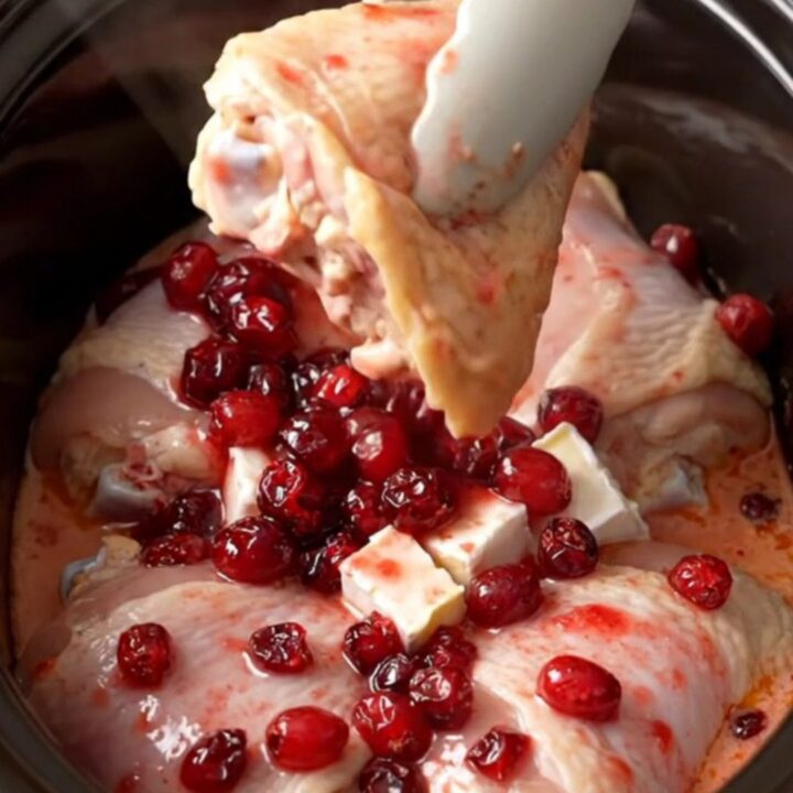A close-up of tender chicken thighs covered in a creamy, pink cranberry and brie sauce inside a slow cooker.