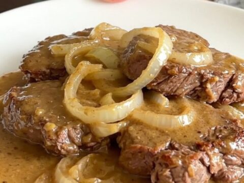 A top-down view of raw cube steaks arranged in a black slow cooker with sliced onions and soup.