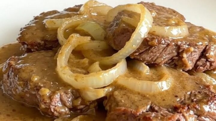 A top-down view of raw cube steaks arranged in a black slow cooker with sliced onions and soup.