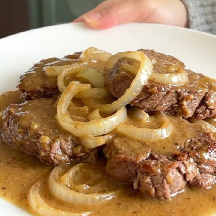 A top-down view of raw cube steaks arranged in a black slow cooker with sliced onions and soup.