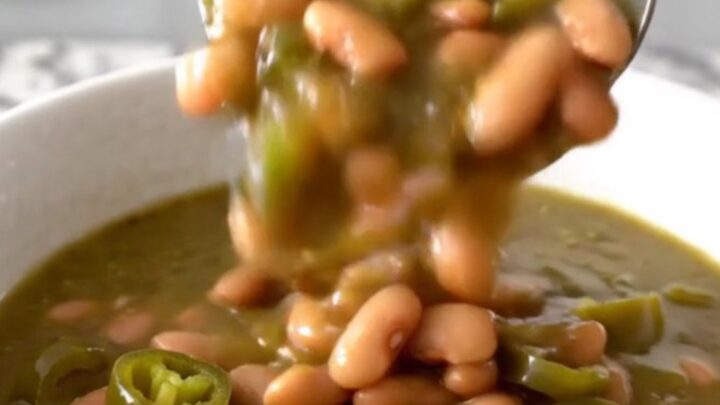A close-up of a white bowl filled with a thick, green-hued pinto bean stew with steam rising gently.