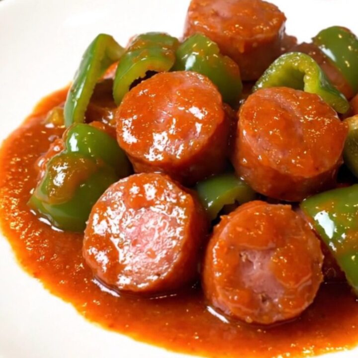 A close-up of honey sriracha kielbasa coins and green peppers in a shiny, red sauce in a slow cooker.