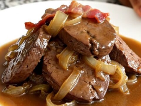A close-up of tender, braised beef liver slices smothered in onions, bacon, and rich brown gravy in a slow cooker.