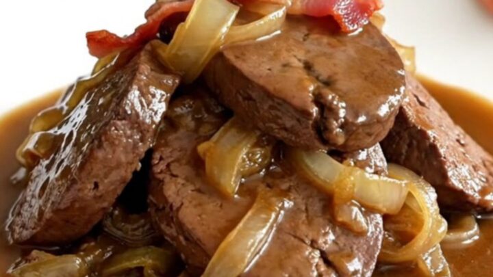 A close-up of tender, braised beef liver slices smothered in onions, bacon, and rich brown gravy in a slow cooker.