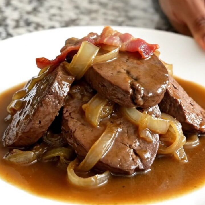 A close-up of tender, braised beef liver slices smothered in onions, bacon, and rich brown gravy in a slow cooker.